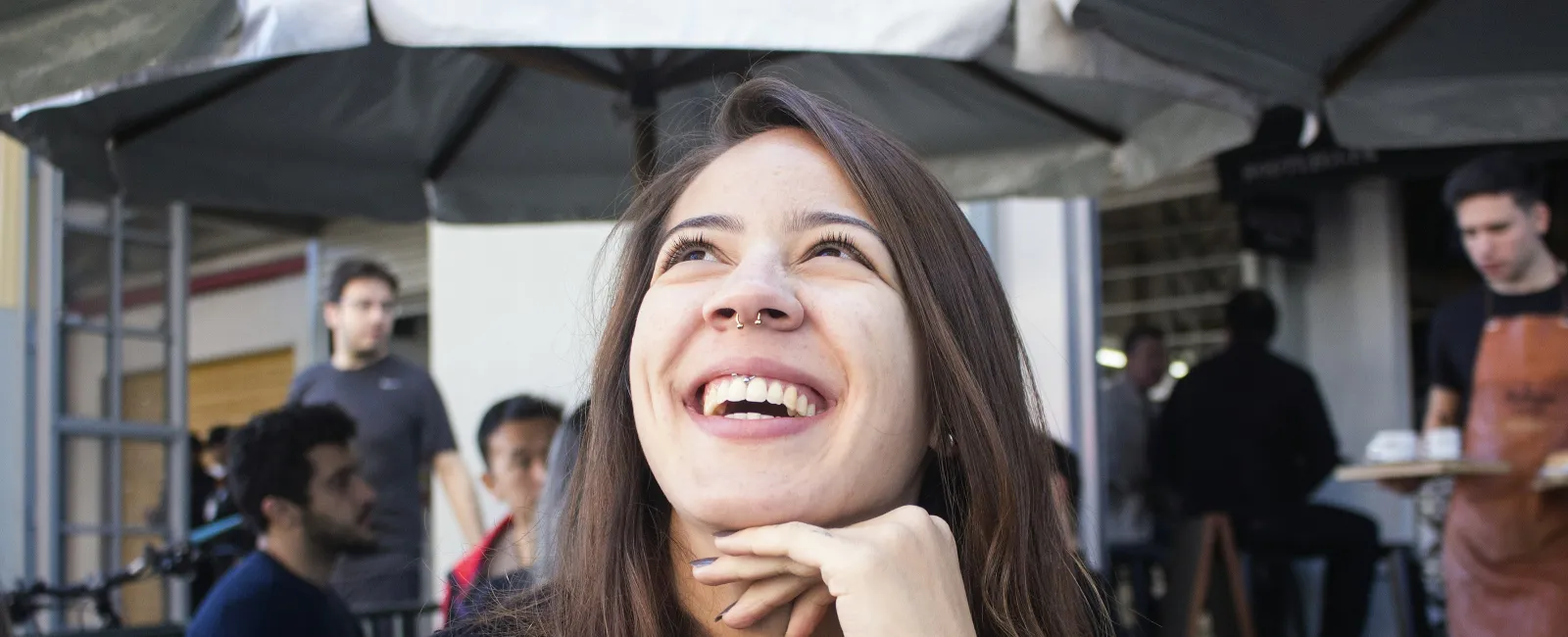 Young woman with nose ring and tattoo laughing outdoors at a café with people and waiter in background