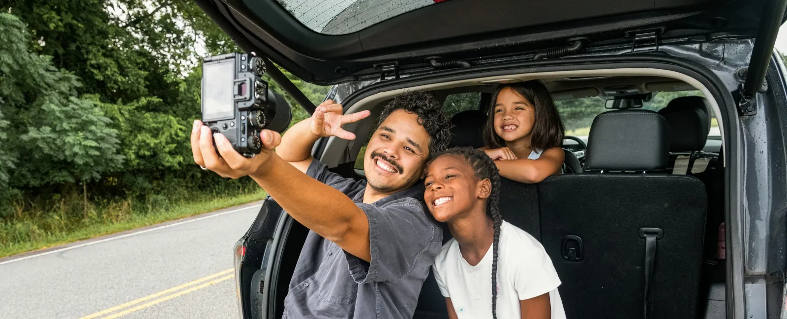 A man and two girls smiling and taking a selfie from the open trunk of a car on a roadside.