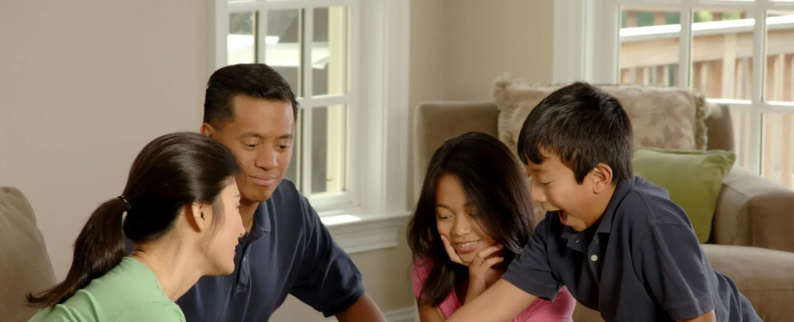 Family of four smiling and playing a board game together around a wooden table in a bright living room