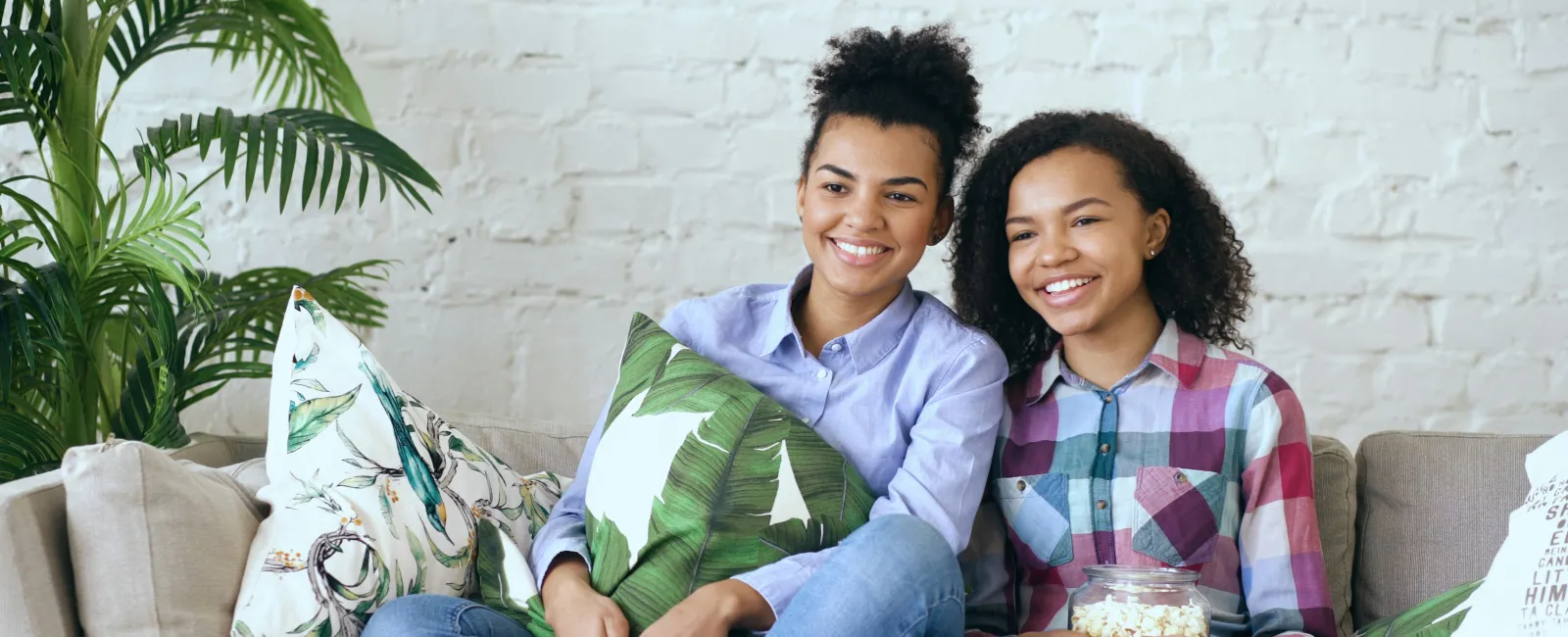 Two young women sitting on a couch enjoying a movie, smiling and holding popcorn and a remote control.