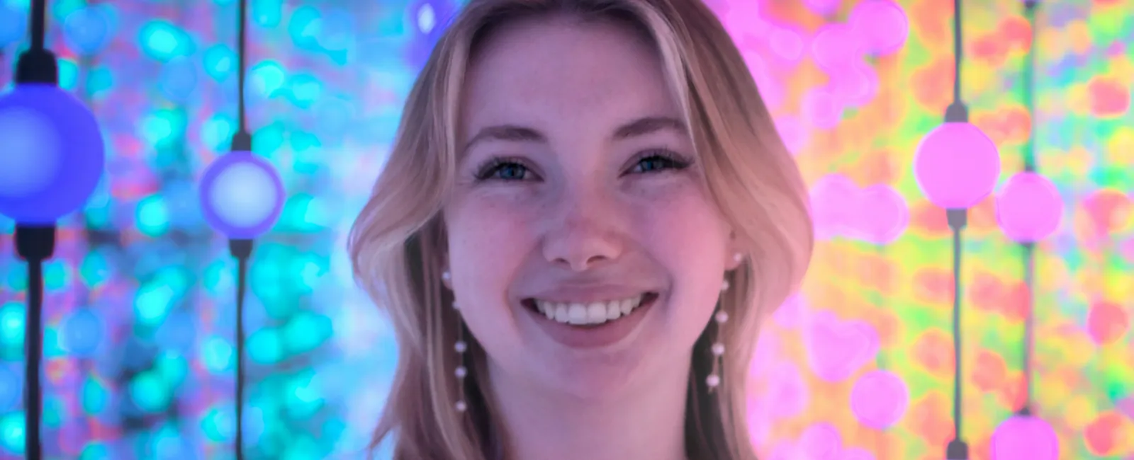 Smiling young woman with blonde hair in front of colorful glowing orbs and vibrant blurred background.
