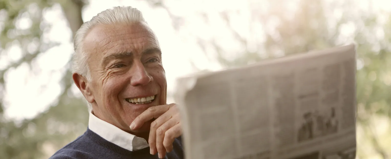 Smiling elderly man enjoying reading a newspaper outdoors with blurred natural background