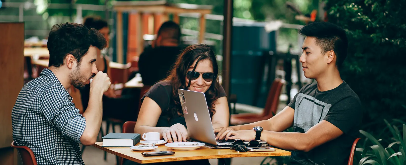 Three people collaborating outdoors at a cafe table with a laptop, coffee, and books in a green garden setting.