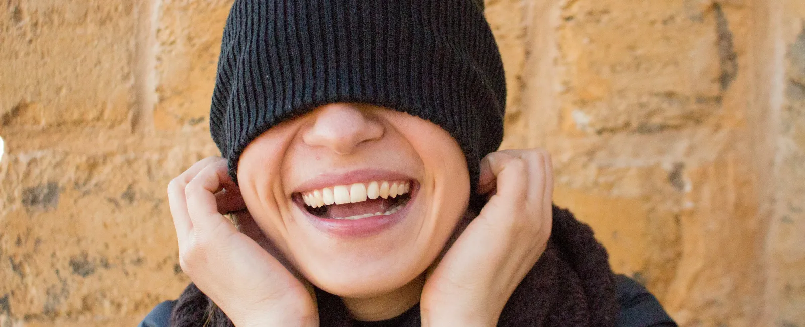 Smiling person with black beanie pulled over eyes laughing against a beige stone wall.