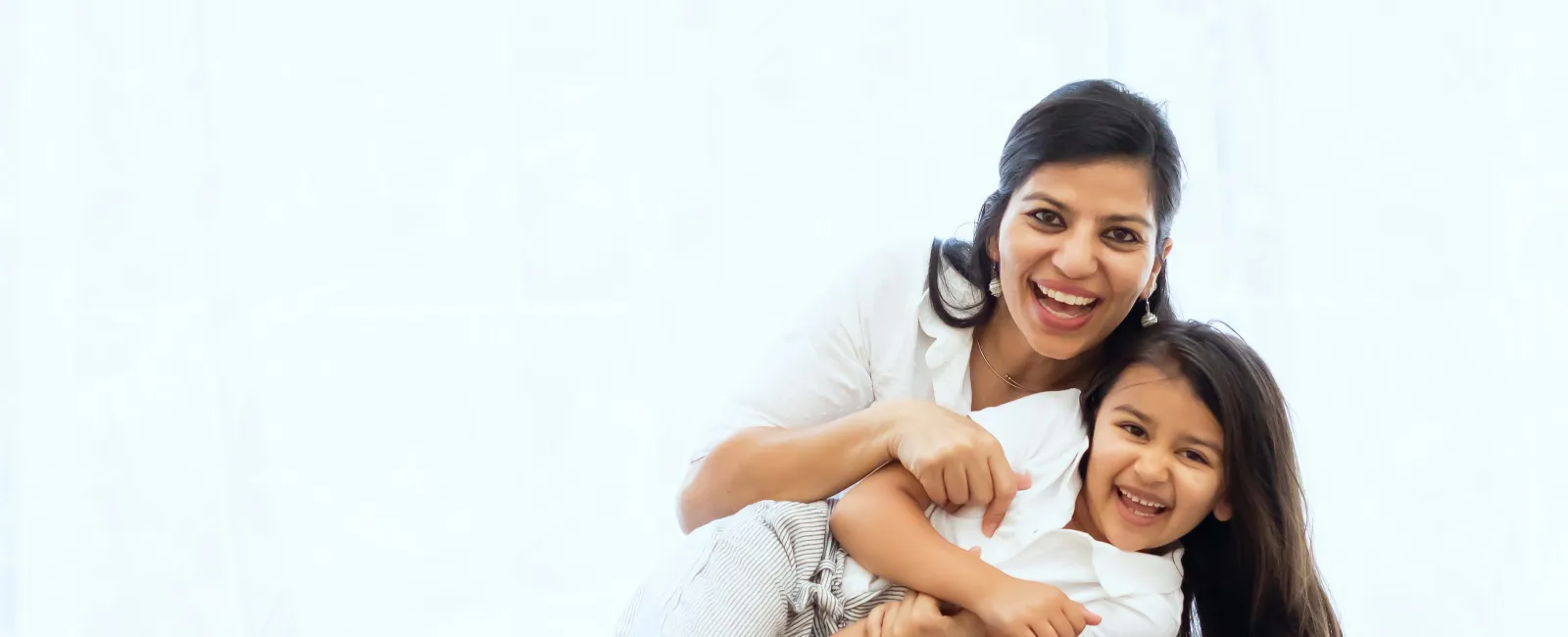 Smiling mother hugging her happy daughter while sitting on a couch in a bright living room.