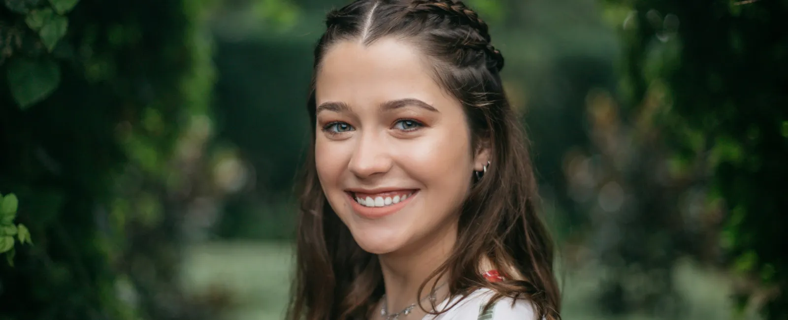 Smiling young woman with braided hair wearing floral dress stands outdoors framed by green foliage.