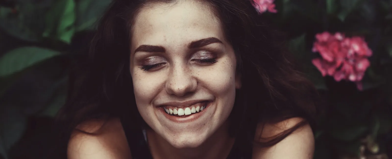 Smiling young woman with closed eyes surrounded by pink and white flowers in a natural outdoor setting