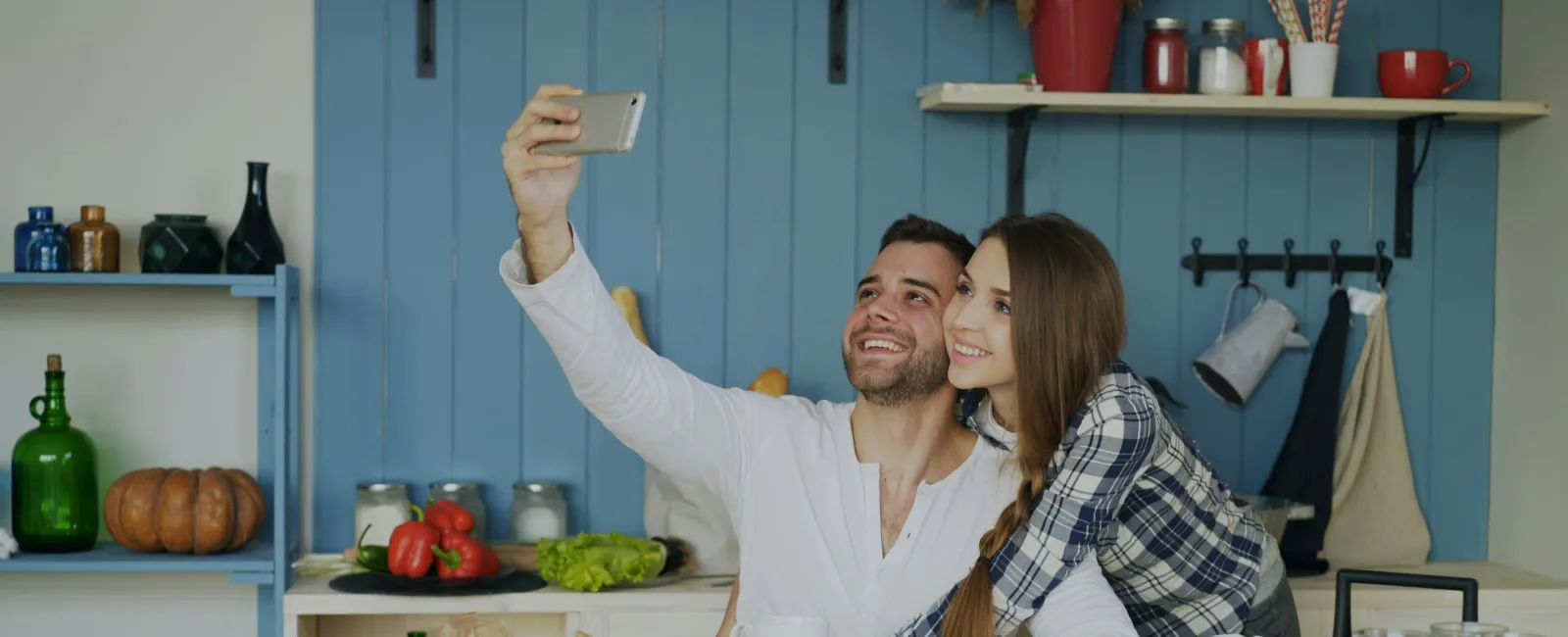 Happy couple taking a selfie in a cozy kitchen with fresh fruits, croissants, and coffee on the table.