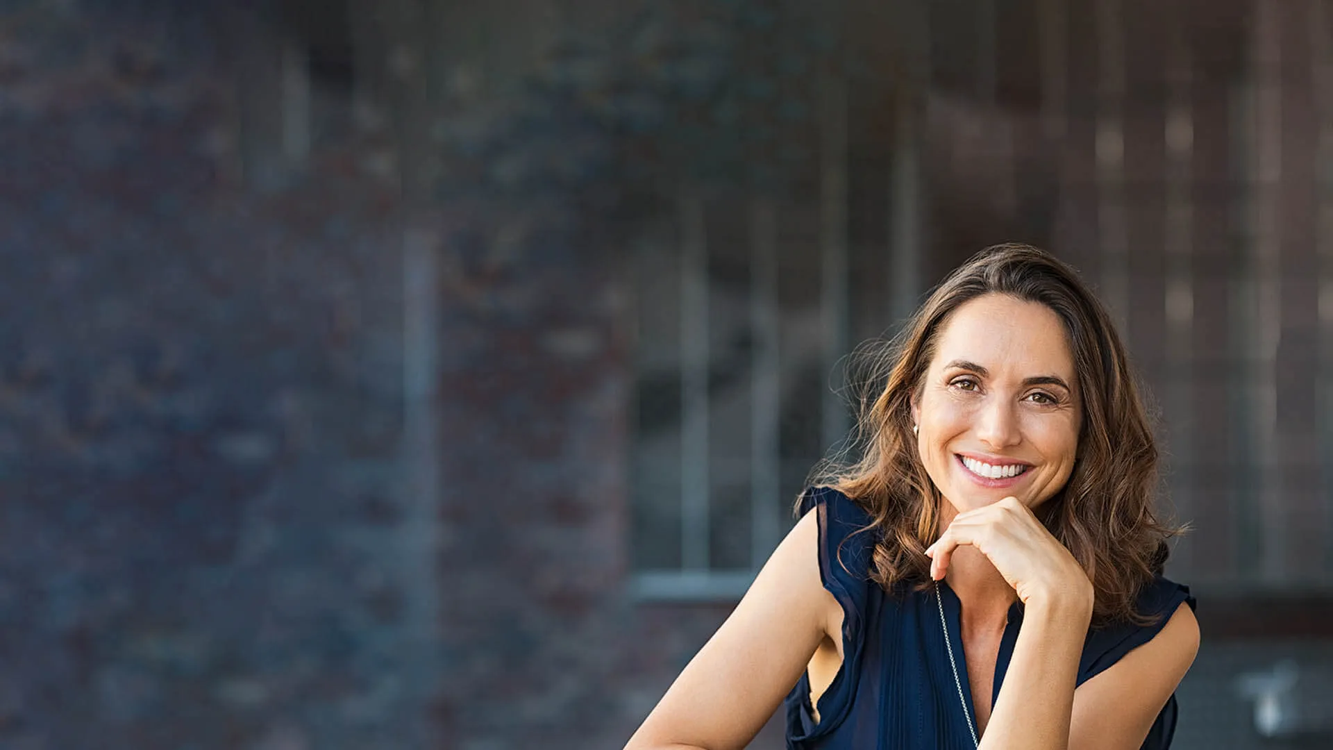 Smiling woman with brown hair in a dark sleeveless top posing with hand on chin against blurred brick background