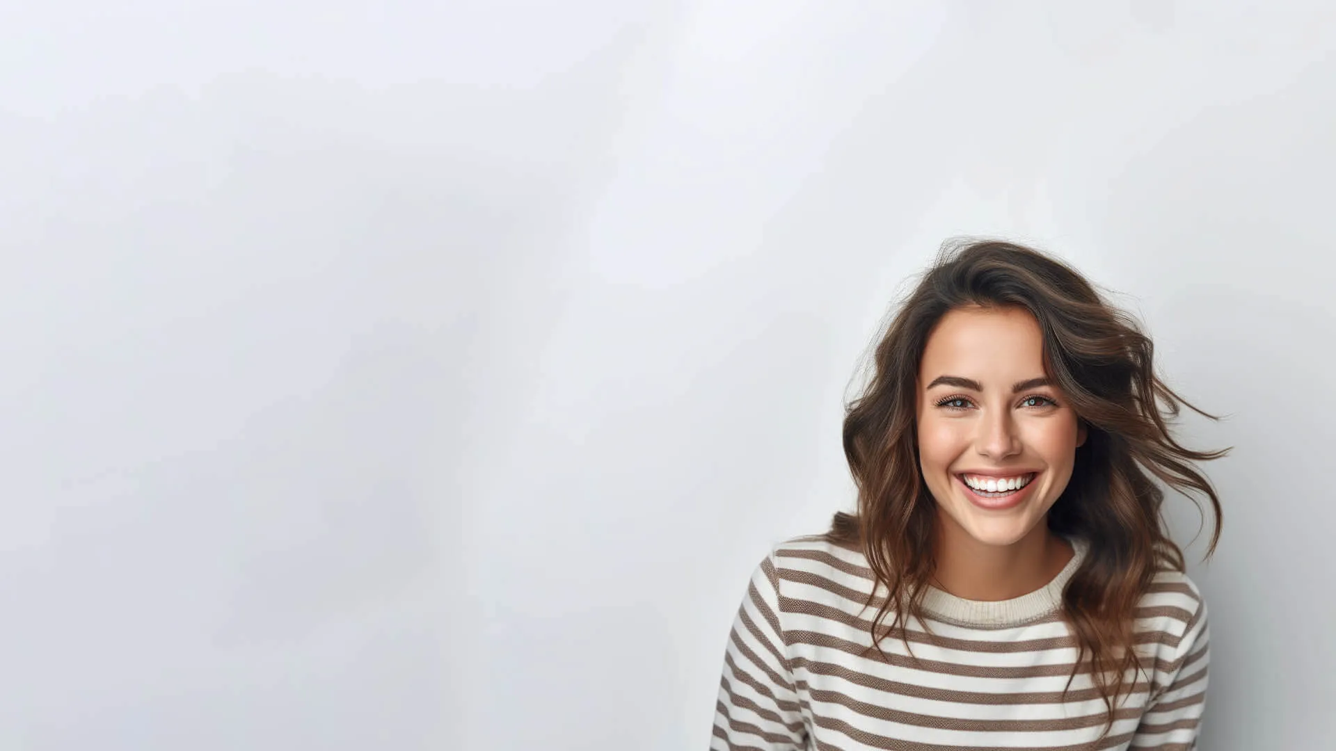 Smiling young woman with wavy hair wearing a striped sweater, standing against a light gray background.