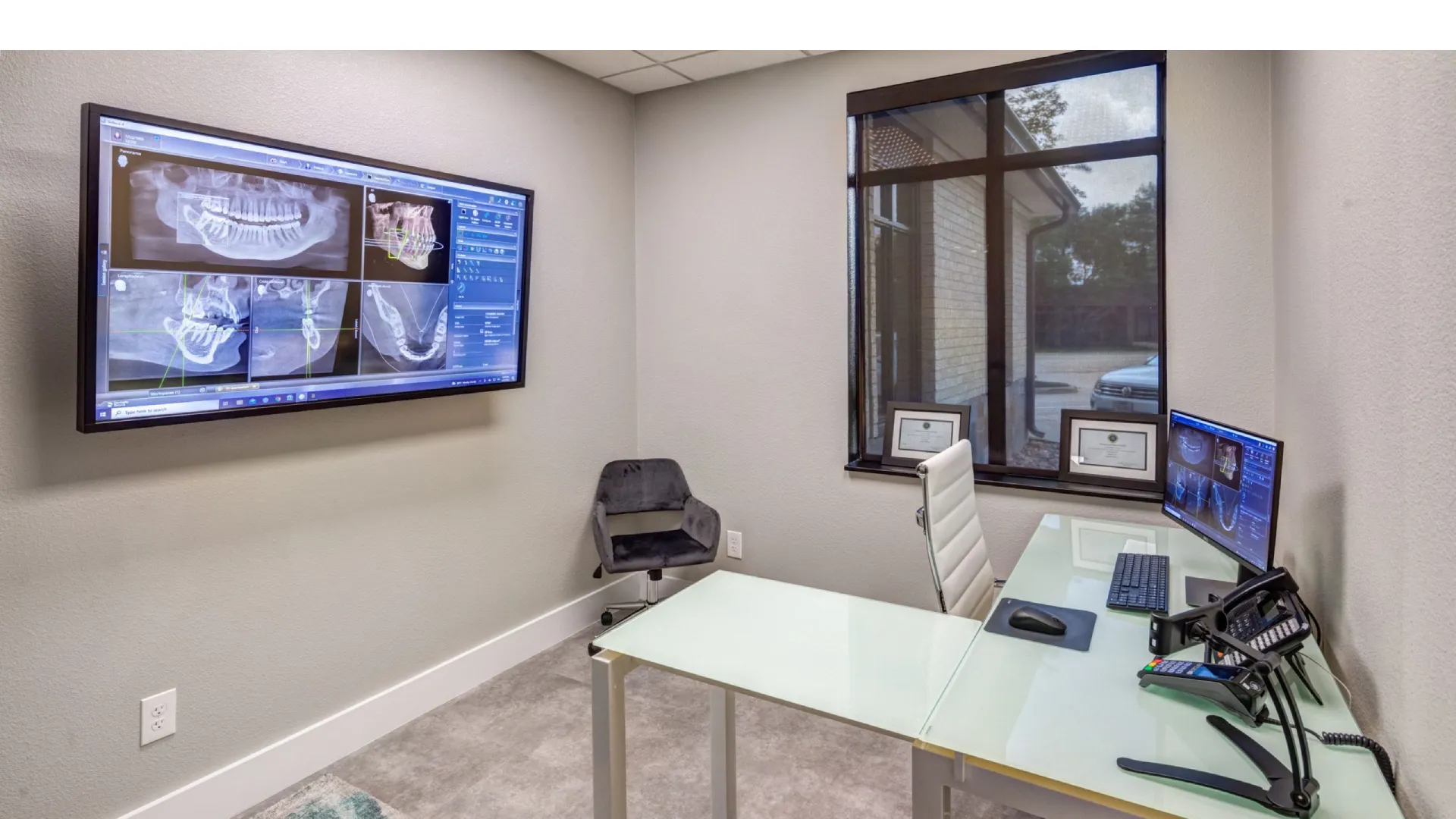Modern dental office with glass desk, computer displaying X-rays, wall-mounted screen showing dental images.