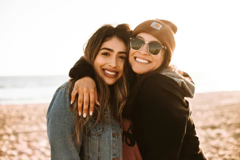 Two women smiling and hugging on a sunny beach, one wearing sunglasses and a beanie, the other a denim jacket.