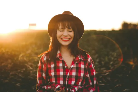 Young woman in a red plaid shirt and black hat smiling with sunset and field background.