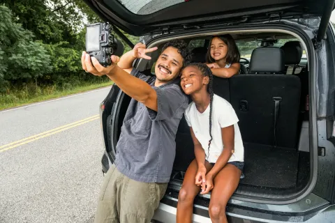 A man and two girls smiling and taking a selfie from the open trunk of a car on a roadside.