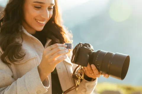 Smiling woman inserting a memory card into a professional camera outdoors during daylight.