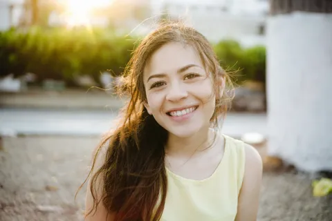 Smiling young woman with long brown hair wearing a yellow sleeveless top outdoors with sunlight in background