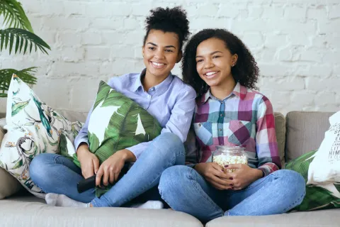 Two young women sitting on a couch enjoying a movie, smiling and holding popcorn and a remote control.