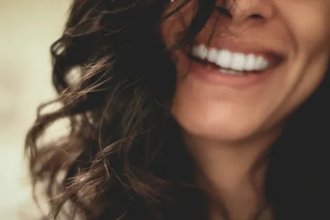 Close-up of a woman smiling showing her white teeth and curly dark hair in natural light.
