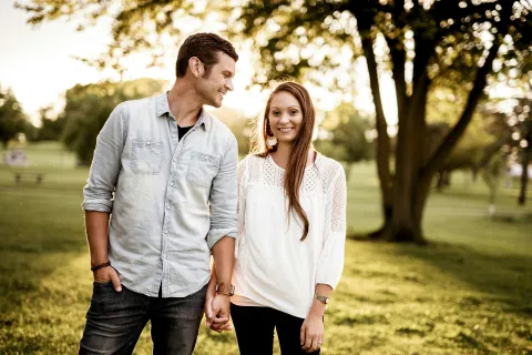 Young couple holding hands and smiling in a sunlit park with trees and green grass in the background
