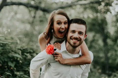 Happy couple outdoors with woman playfully hugging man from behind holding a red flower surrounded by greenery