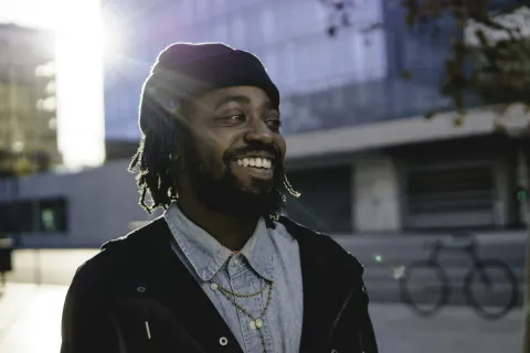 Smiling man with dreadlocks wearing a beanie and layered necklaces outdoors in urban setting with sunlight behind him