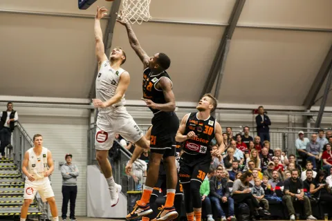 Basketball player in white jumps for a layup while defenders in black jerseys attempt to block the shot at indoor game.
