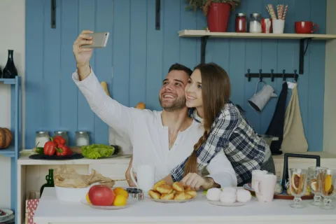 Happy couple taking a selfie in a cozy kitchen with fresh fruits, croissants, and coffee on the table.