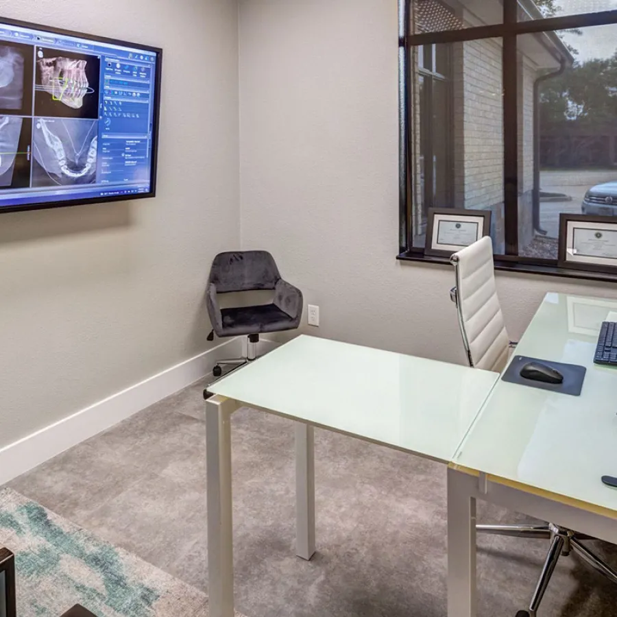 Modern dental office with glass desk, computer monitors displaying X-ray images, and ergonomic chairs in a bright room.