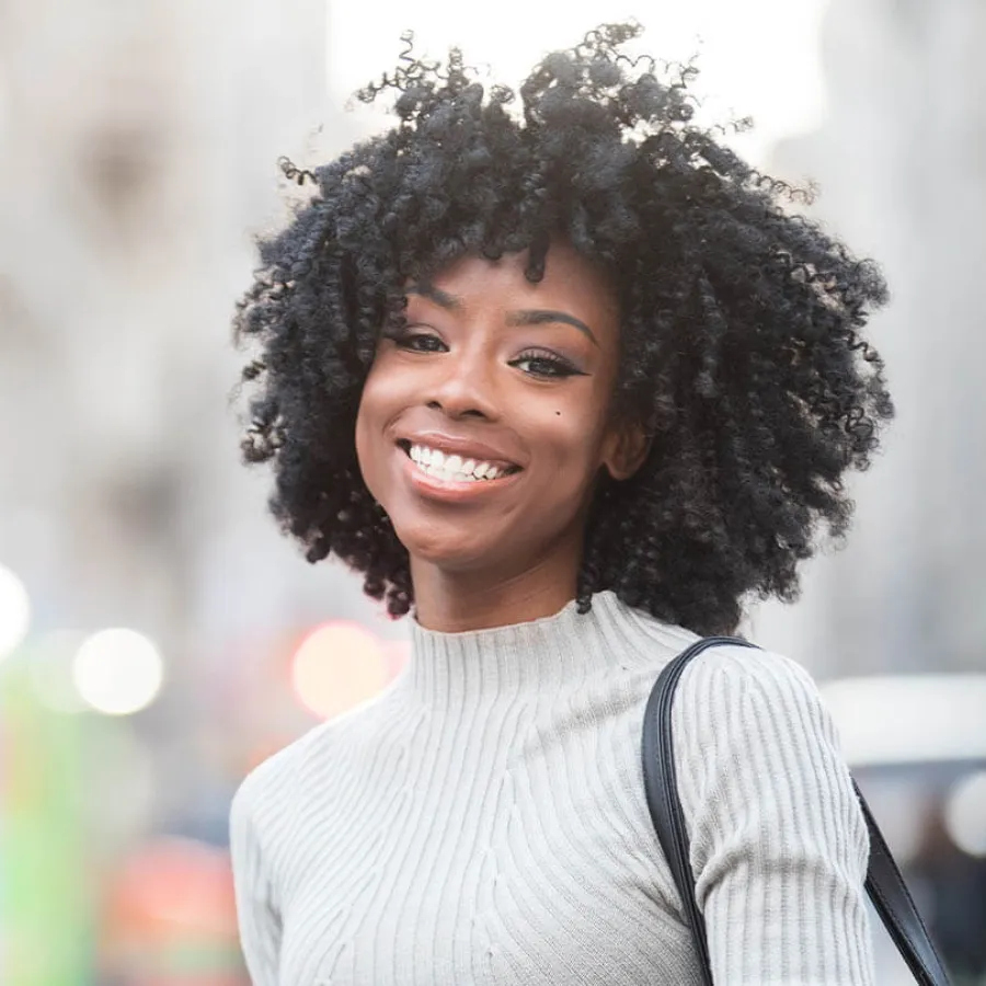 Smiling woman with natural curly hair wearing a light ribbed sweater and carrying a black shoulder bag outdoors.