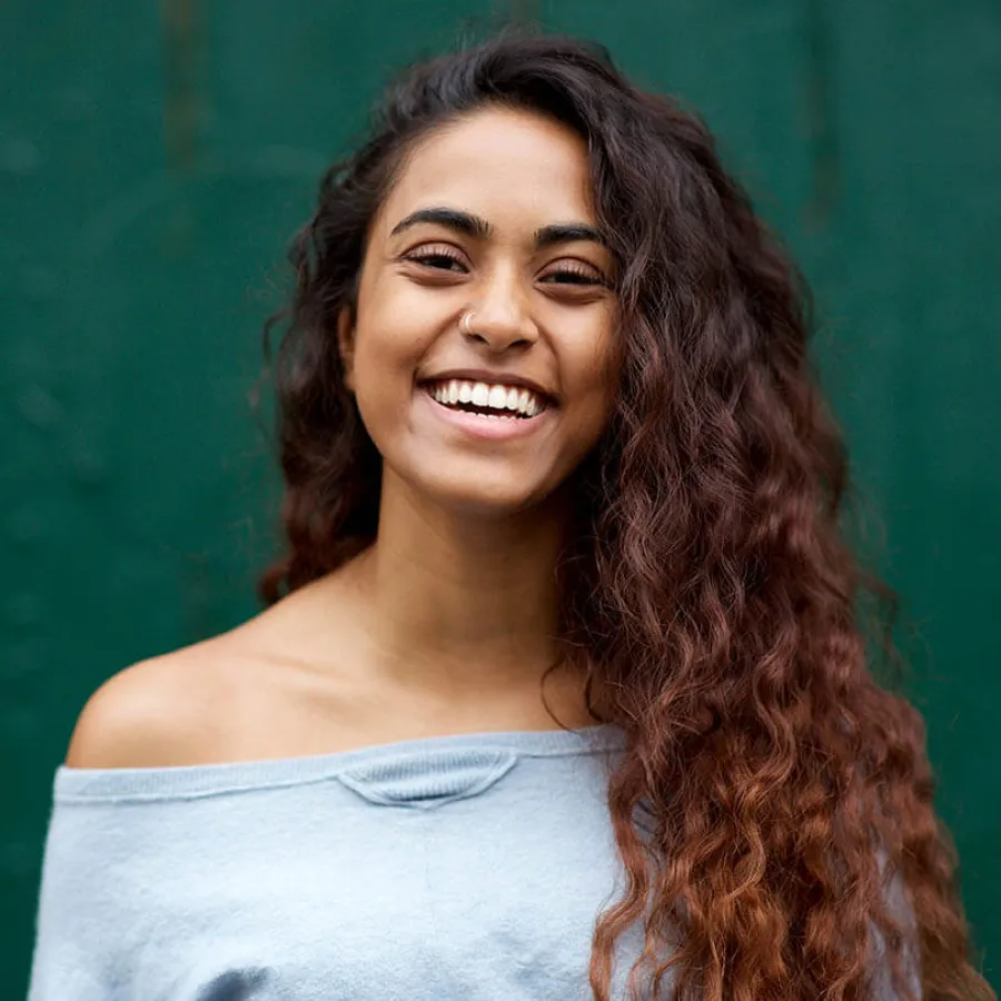 Smiling young woman with long curly hair wearing an off-shoulder light blue top against a green background