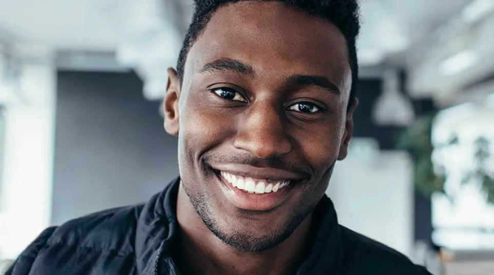 Smiling young African American man wearing a black jacket in a modern indoor setting with natural lighting.
