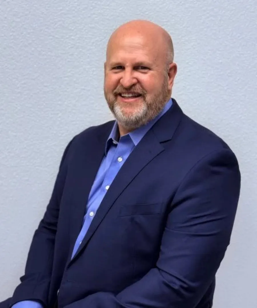 Smiling bald man with beard wearing a navy suit and blue shirt against a plain light background